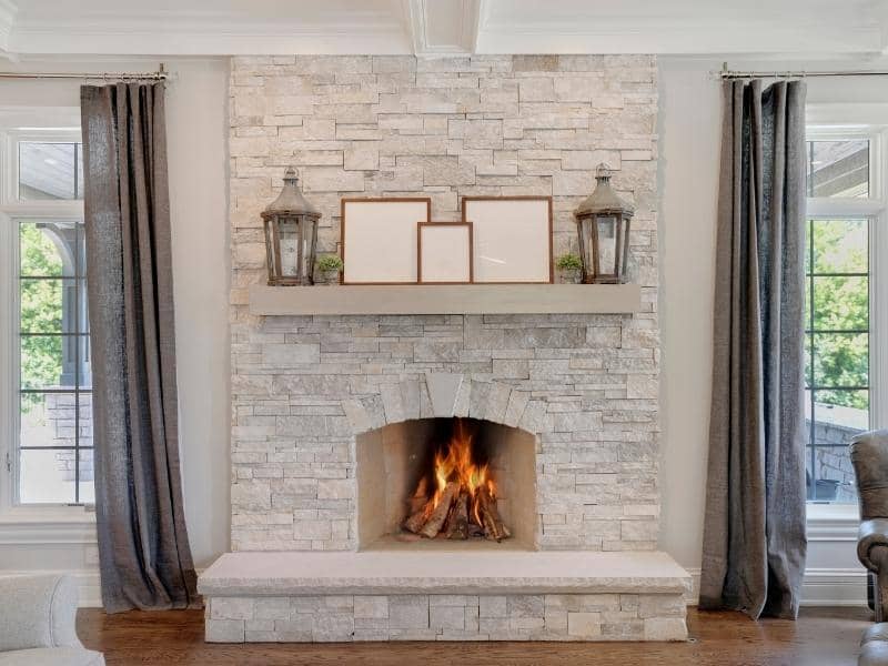Stone fireplace with a crackling fire and a wide stone hearth, flanked by tall windows and dark curtains in a bright living room.