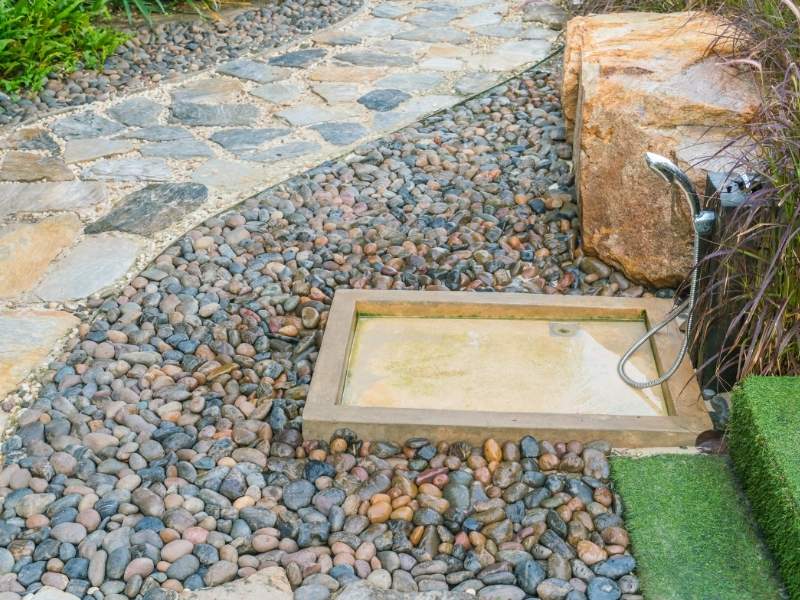 Stone Pavers Walkway Around Outdoor Shower Frisco Texas - Frisco Brick & Stone Outdoor shower with a handheld nozzle beside a rectangular stone basin, surrounded by smooth river rocks and a stone pathway.